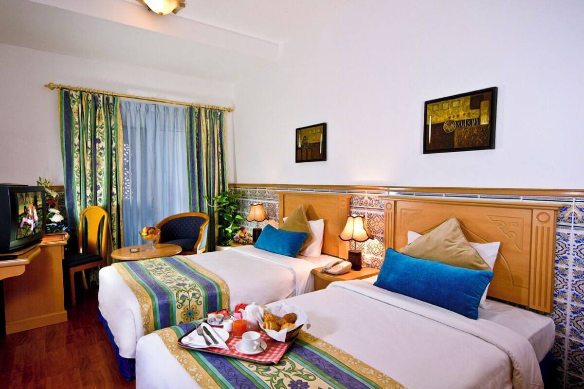 Une chambre d’hôtel avec deux lits, une télévision, une petite table avec le petit-déjeuner, et des coussins décoratifs.