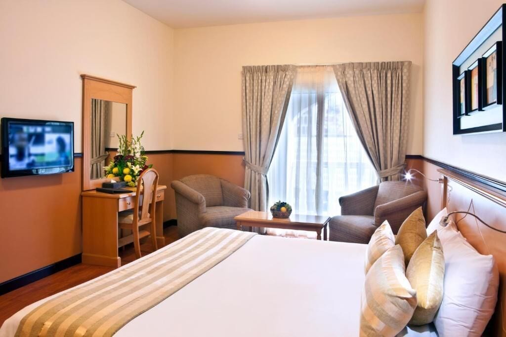 Habitación de hotel con cama, dos sillas, escritorio, televisión y ventana con cortinas.