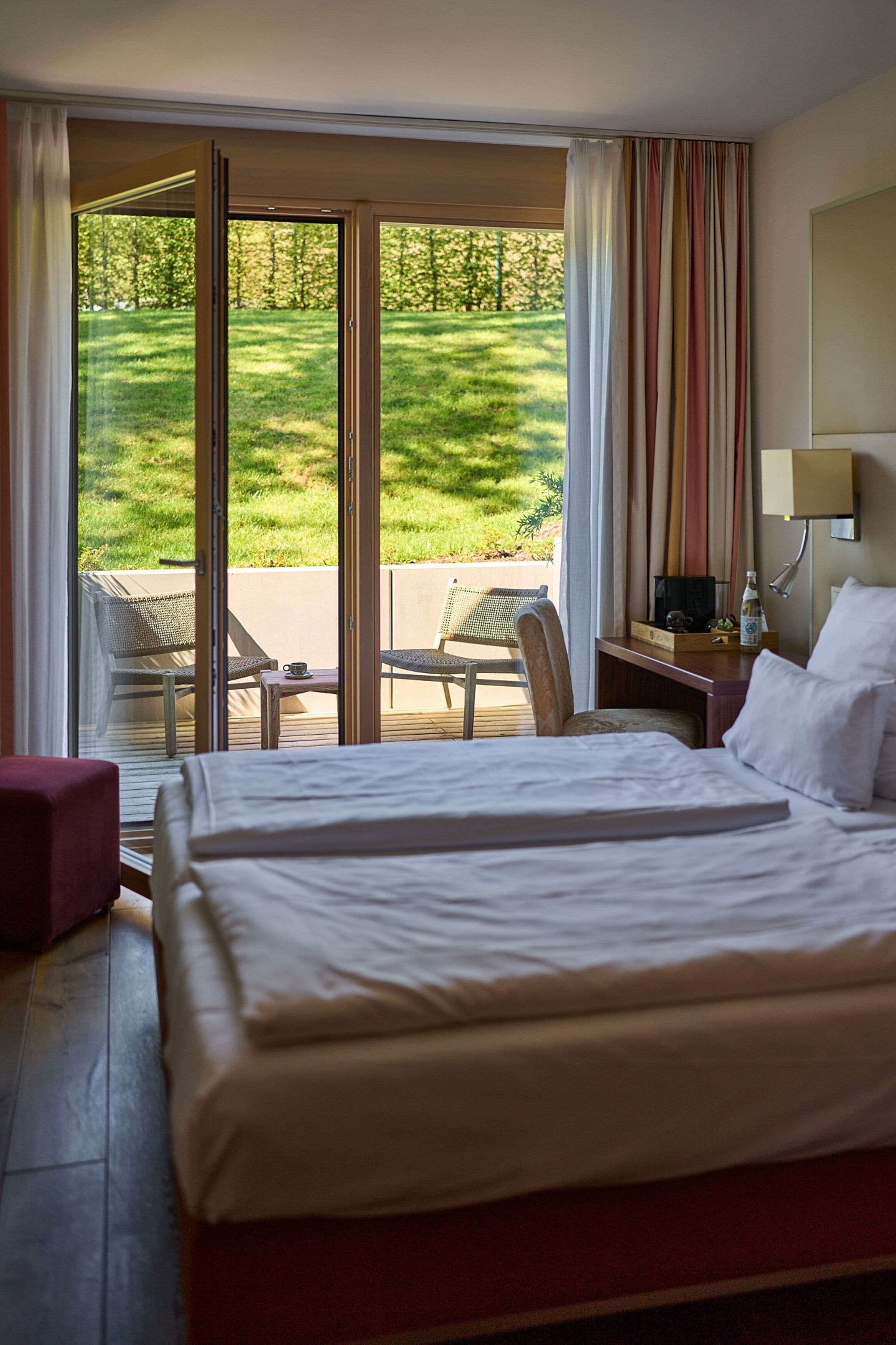A hotel room with a bed, a desk, a chair, and a view of a grassy area through large sliding doors.
