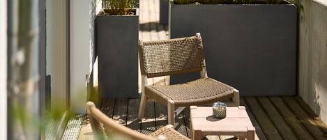A wooden deck with a wicker chair and a small table, surrounded by potted plants and a concrete wall.