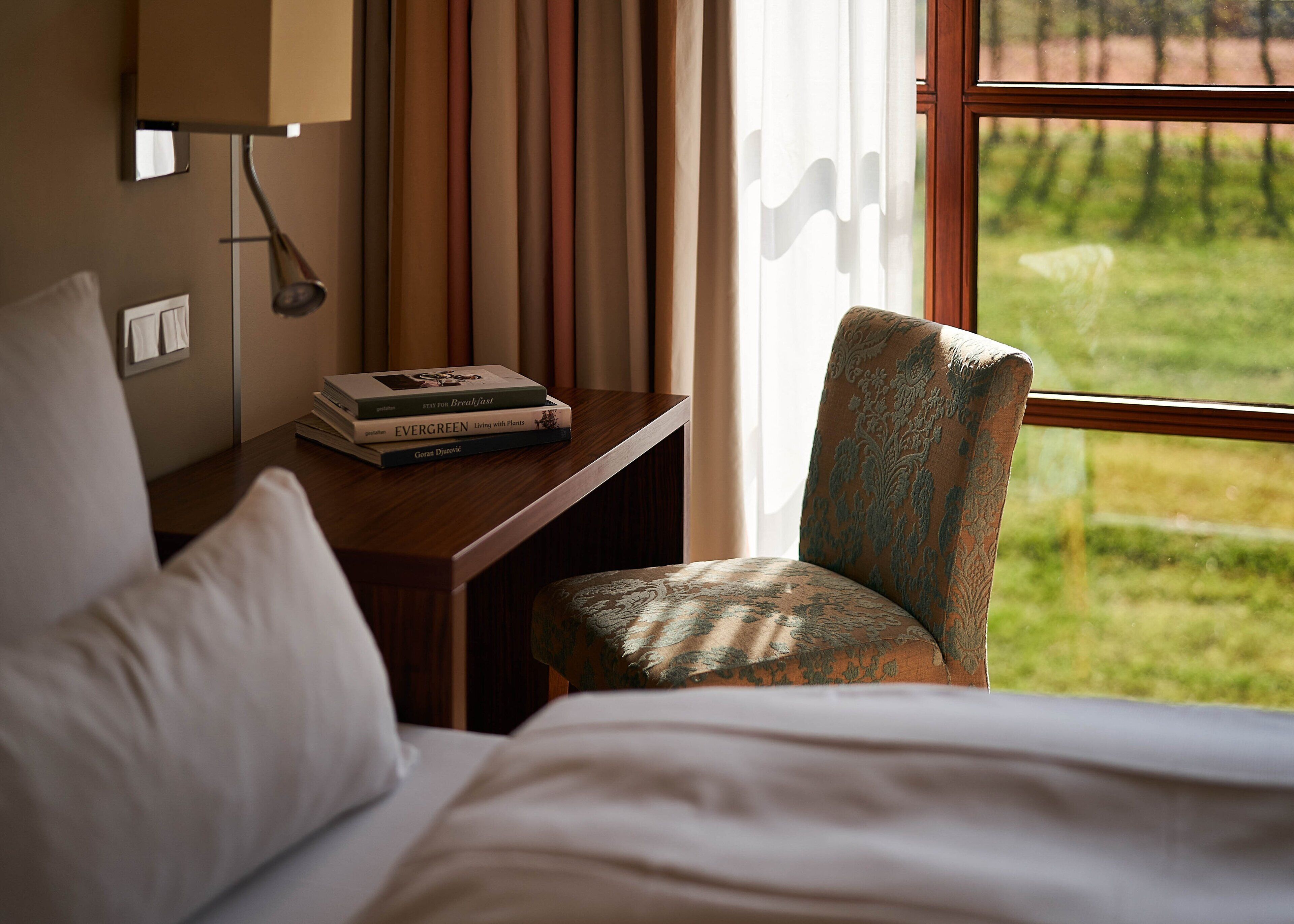 Habitación de hotel con cama, una silla, una mesita con libros, una ventana con vista a follaje y una lámpara fijada a la pared.
