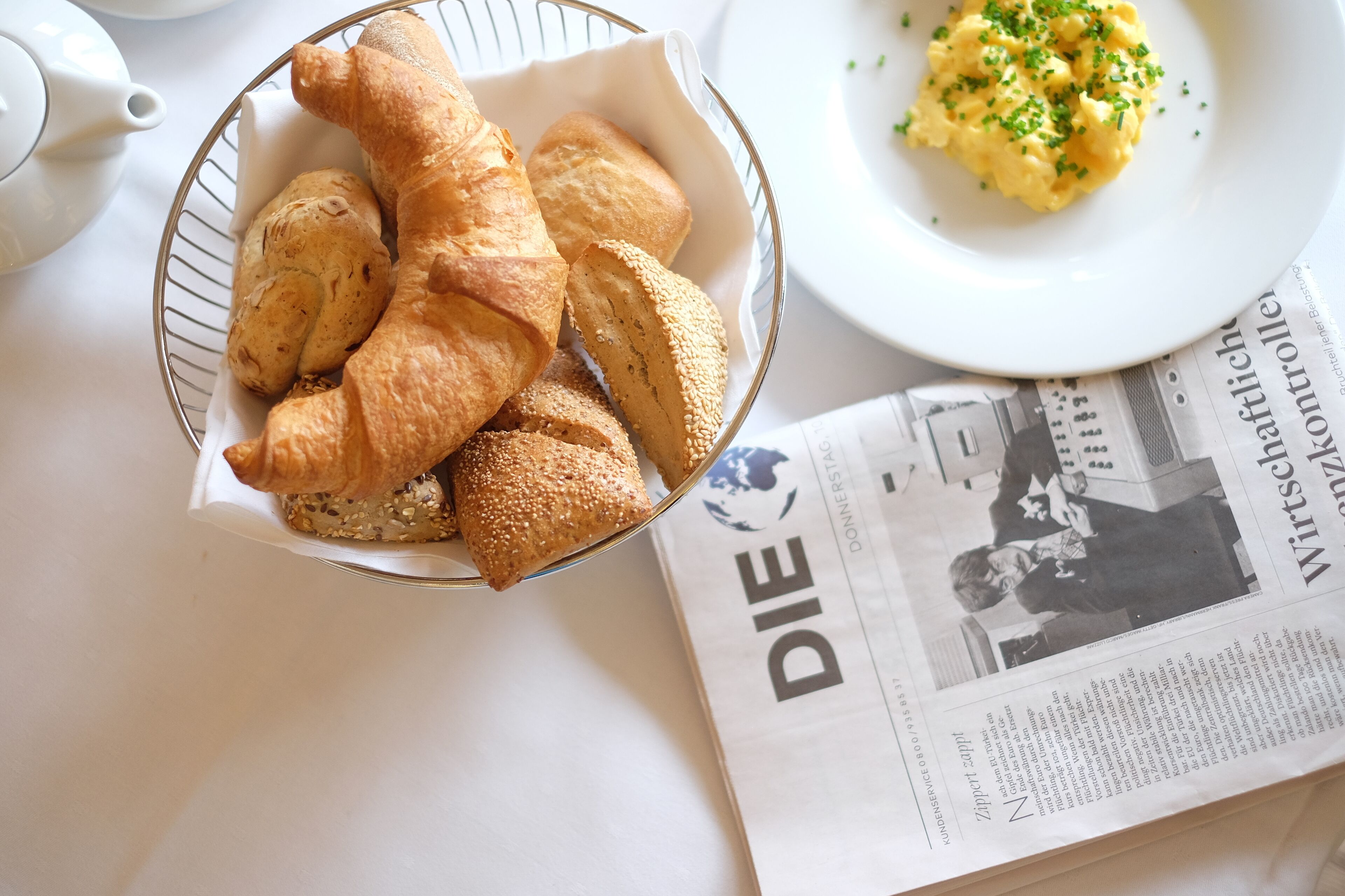 A bowl of croissants and a plate of scrambled eggs on a table with a newspaper.