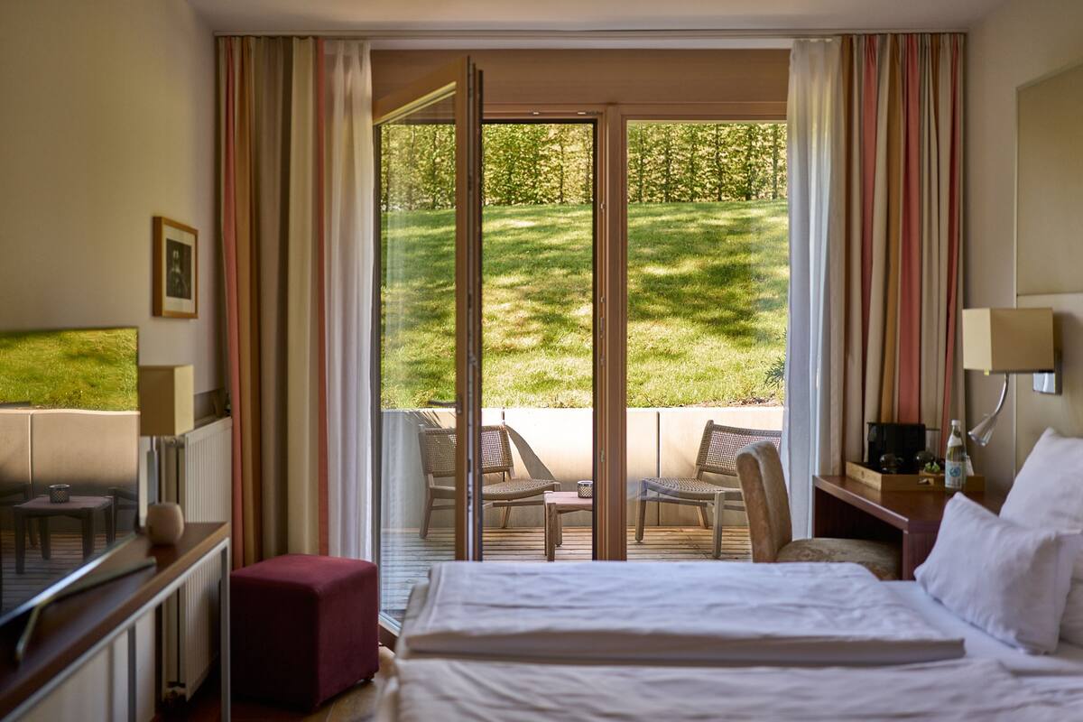 A hotel room with a bed, a desk, a chair, and a view of a grassy outdoor area through a sliding glass door.
