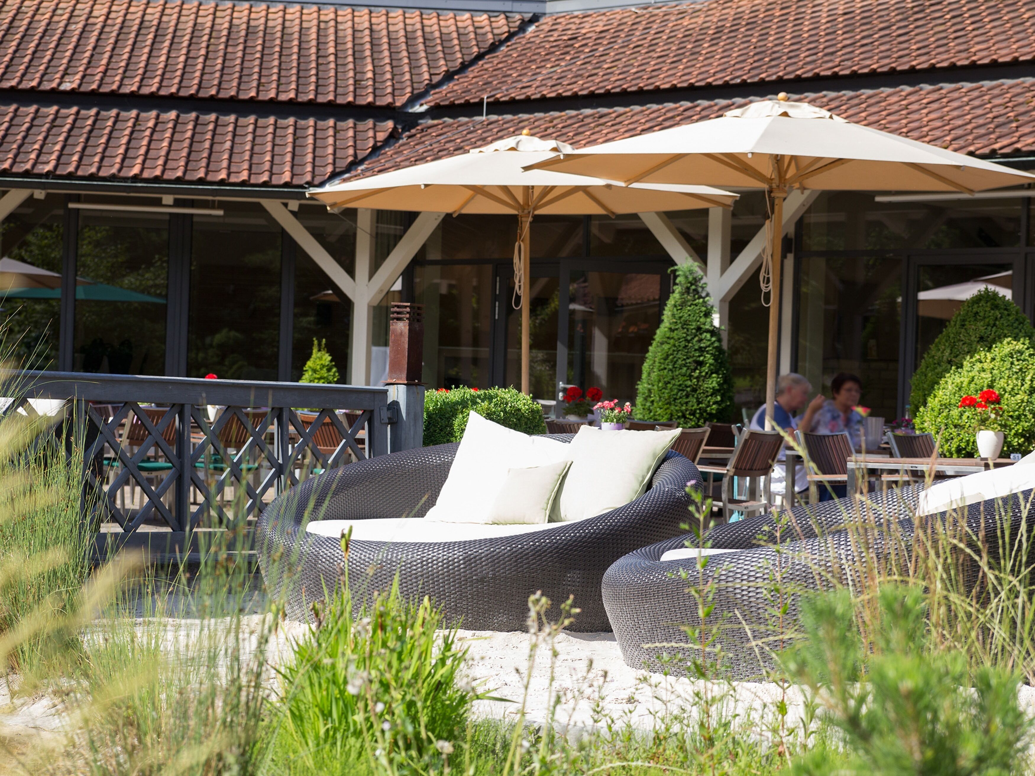 An outdoor seating area with wicker furniture and umbrellas, surrounded by greenery and a building with a tiled roof.