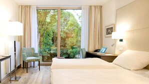 Une chambre d’hôtel avec une grande fenêtre, un lit, un bureau avec une lampe, une chaise et un ordinateur portable.