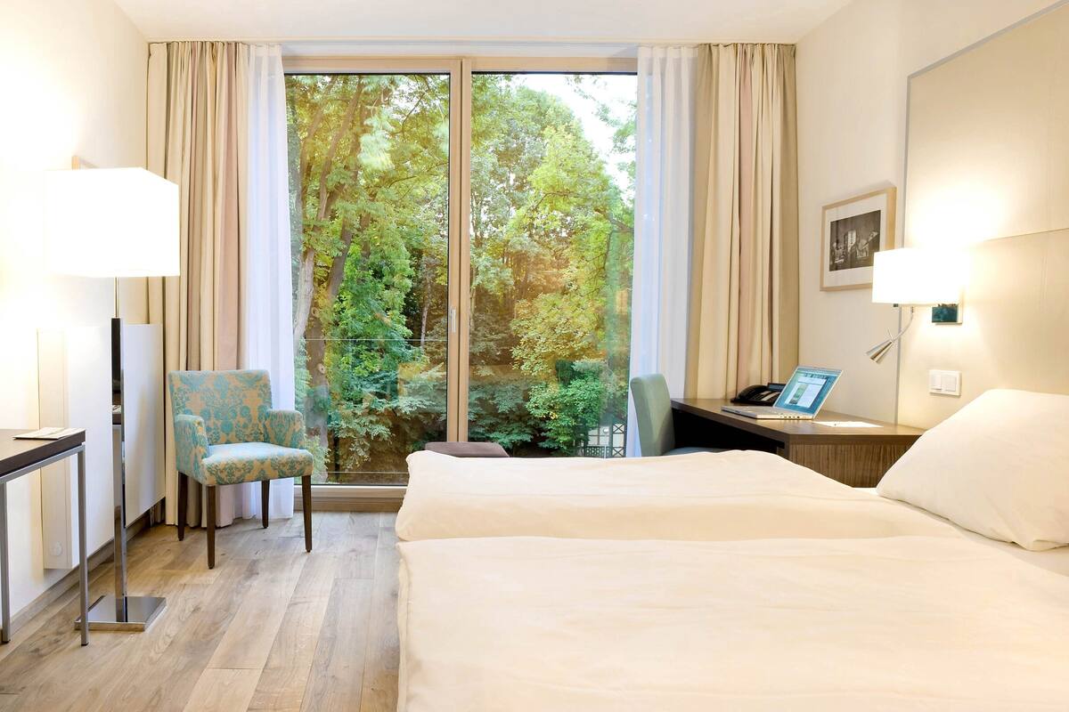 A hotel room with a large window, a bed, a desk with a lamp, a chair, and a laptop.
