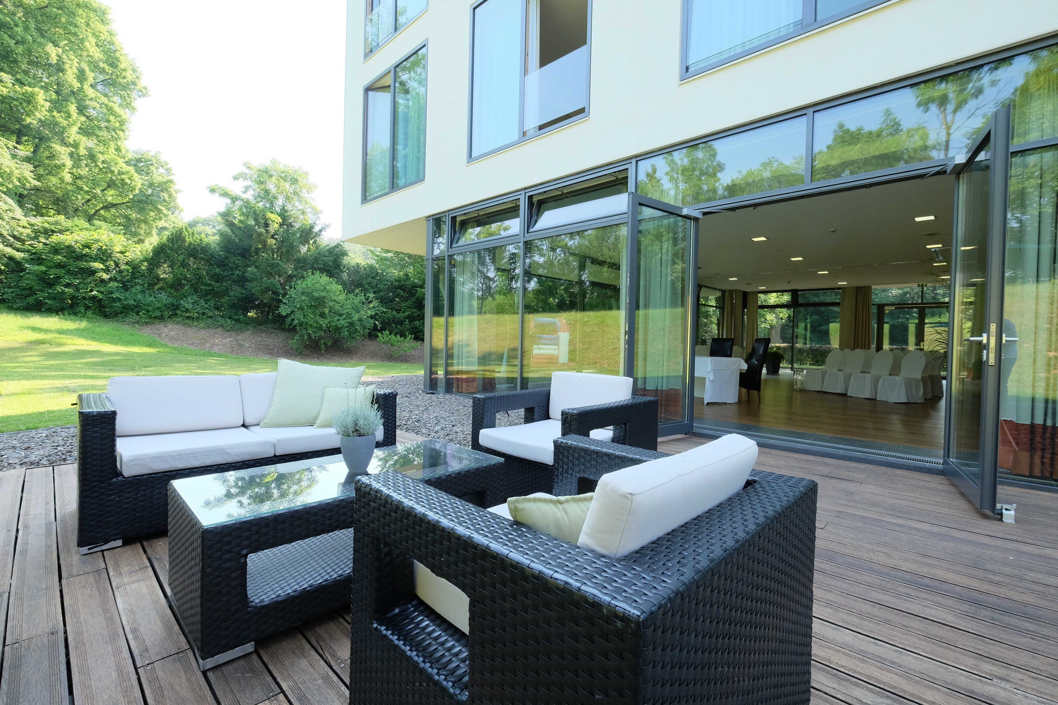 An outdoor patio with wicker furniture, a glass table, and a view of a modern building with large windows.