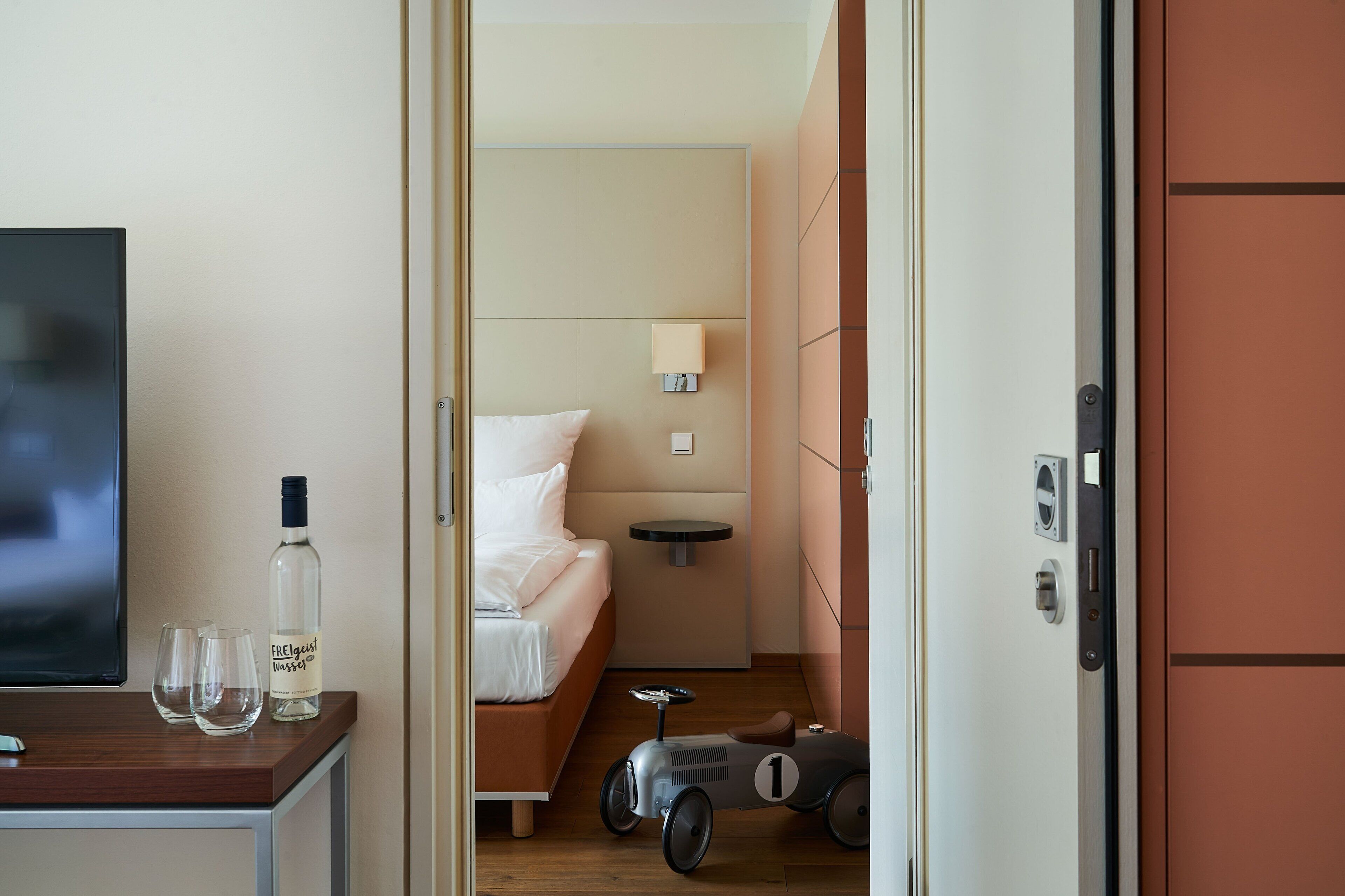 A hotel room with a bed, a small table with a bottle and glasses, a toy car, and a TV on a wooden surface.