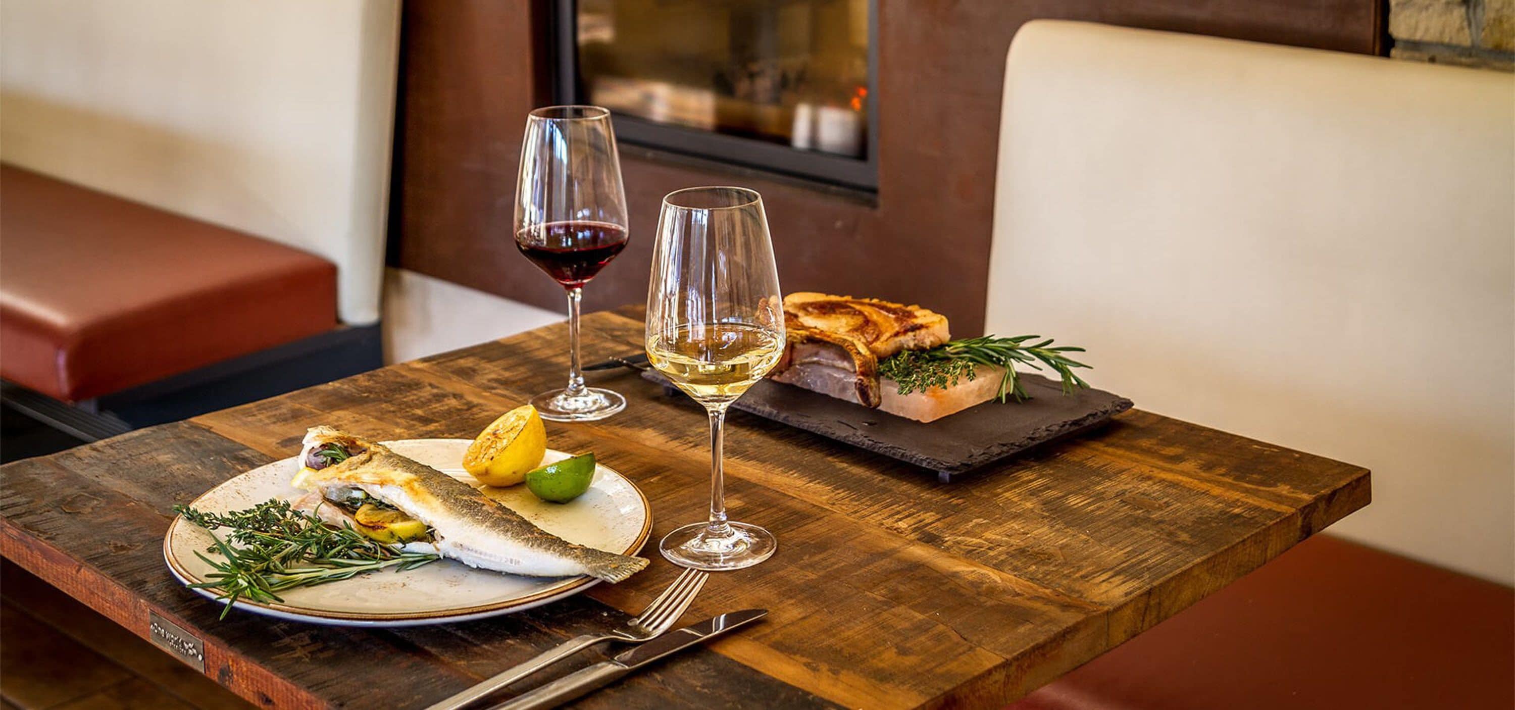 Ein Holztisch mit einem Teller Fisch, Zitrone, Kräutern, zwei Weingläsern und einem Stück Brot auf einer Schieferplatte.