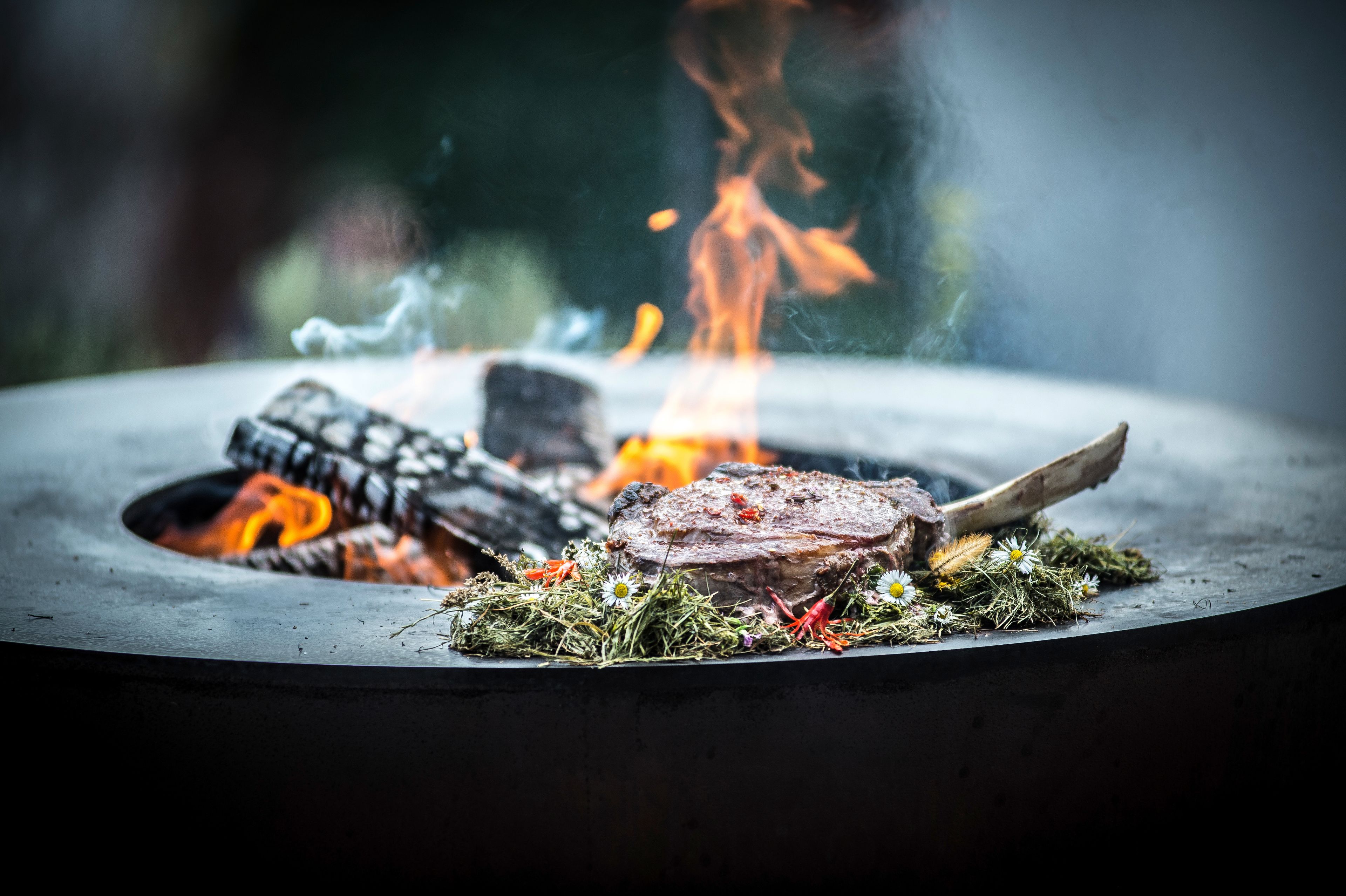 Ein Steak mit Knochen auf einem Grill, umgeben von Flammen, Grünpflanzen und Blumen.