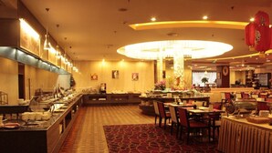 Ampla área de refeições de hotel com serviço de buffet, um candelabro central e piso revestido de carpete vermelho.