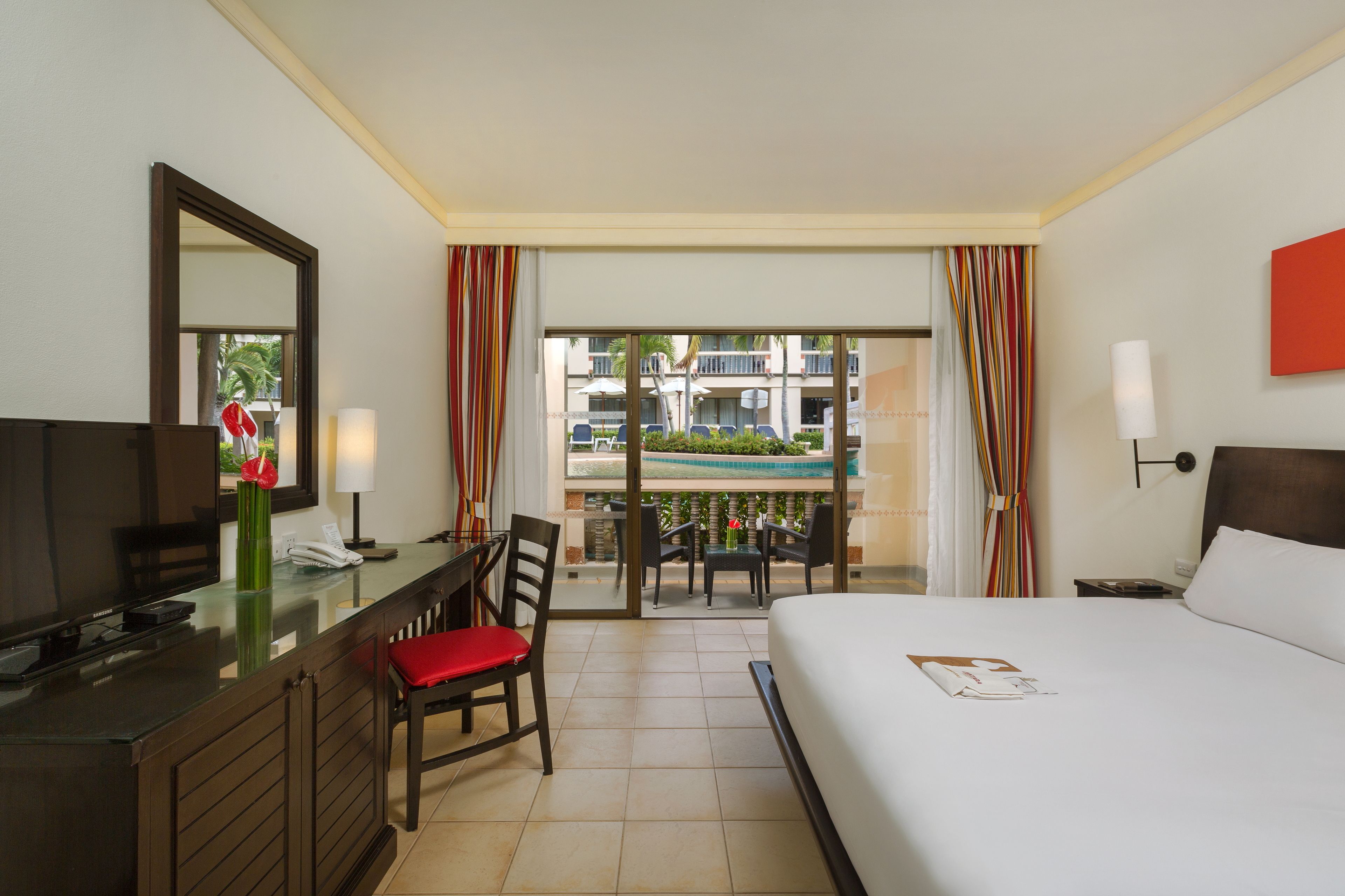 Camera d'albergo con un letto, una scrivania con una sedia, una TV, una finestra con tende e vista su una piscina e edifici esterni.