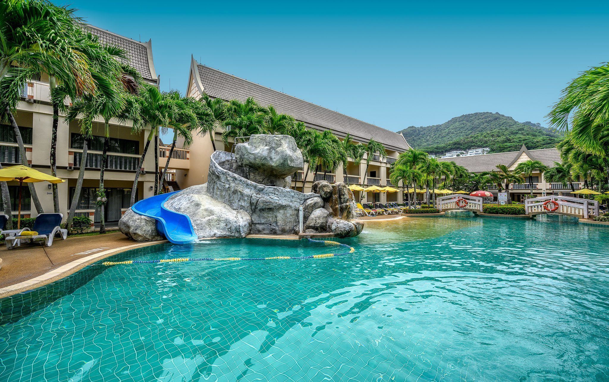 A resort pool with a water slide, palm trees, and a mountainous backdrop.