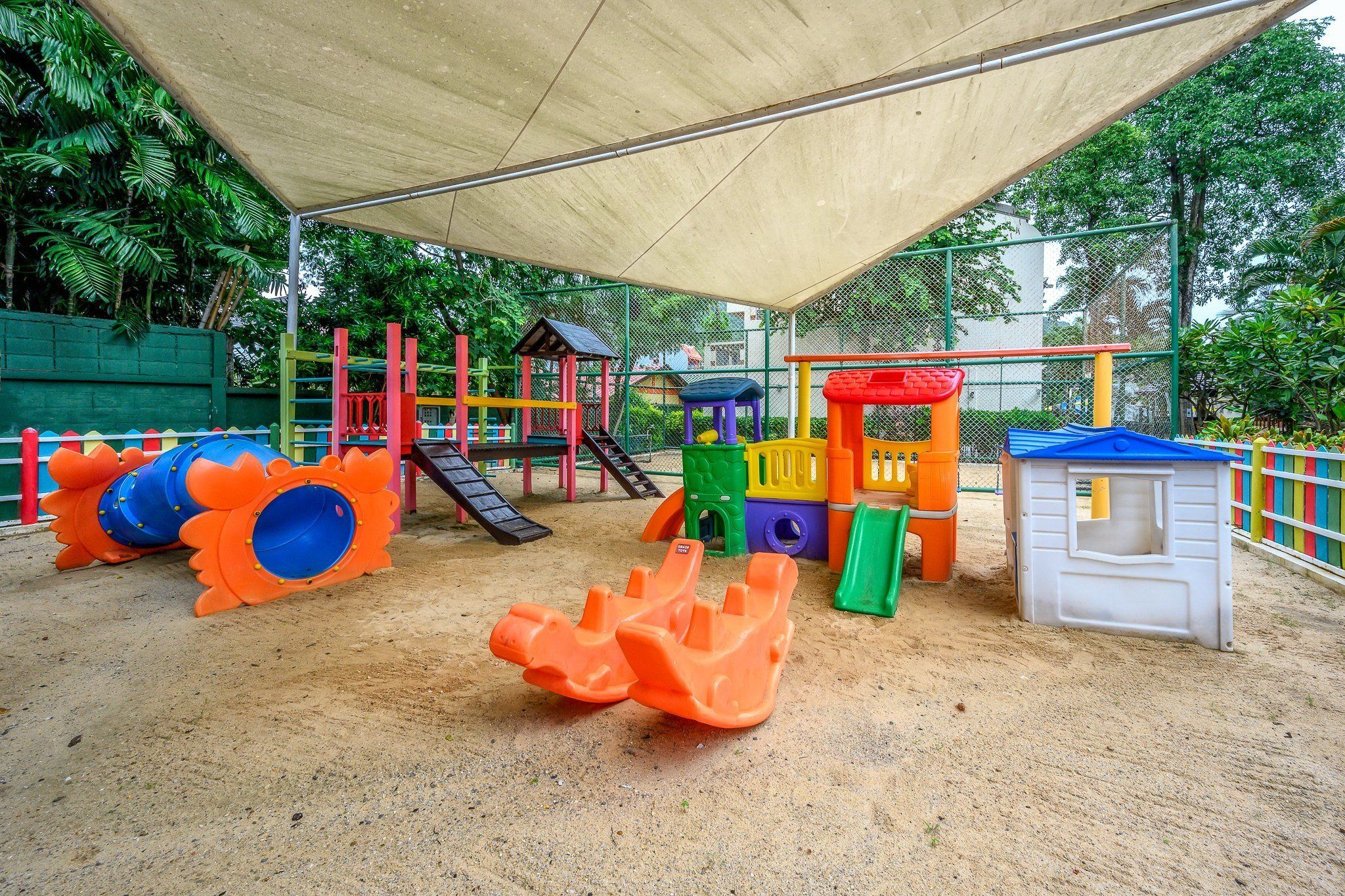 A playground with various play structures including slides and climbing areas, surrounded by a fence and trees.