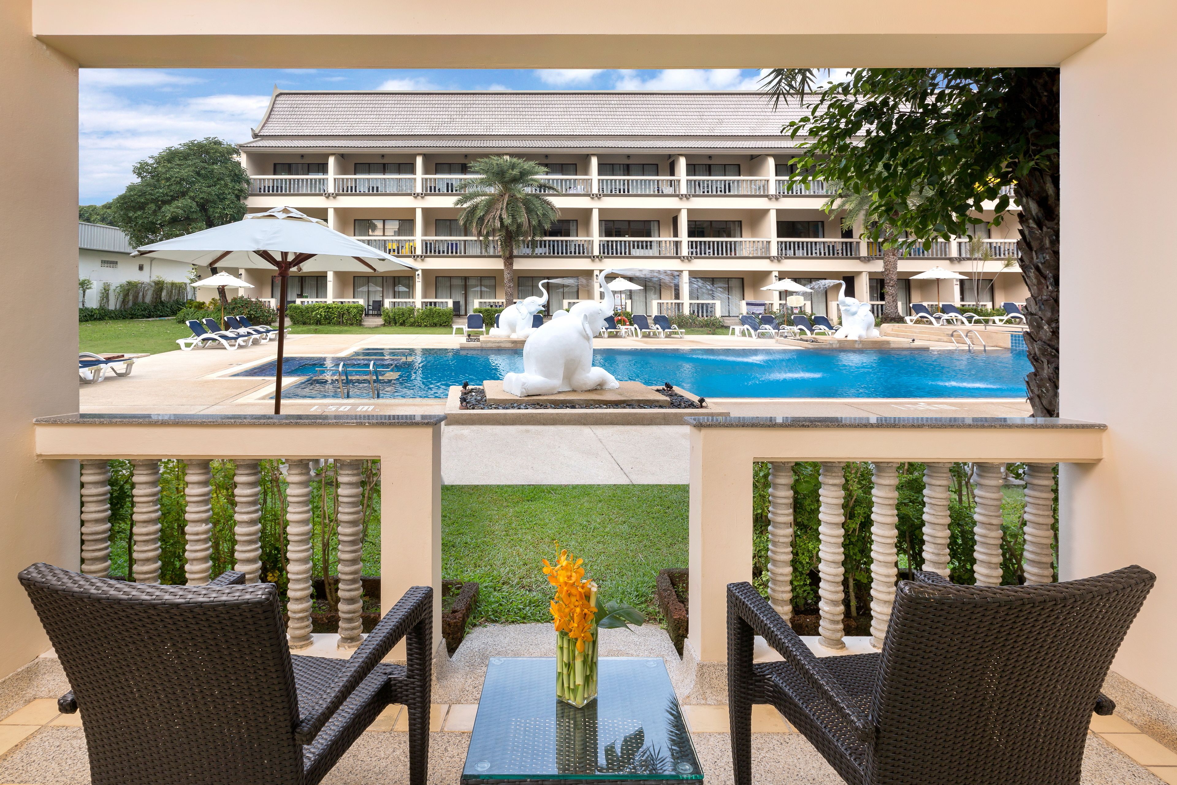 A poolside view with lounge chairs, a table with a vase of flowers, and a building with a fountain in the background.