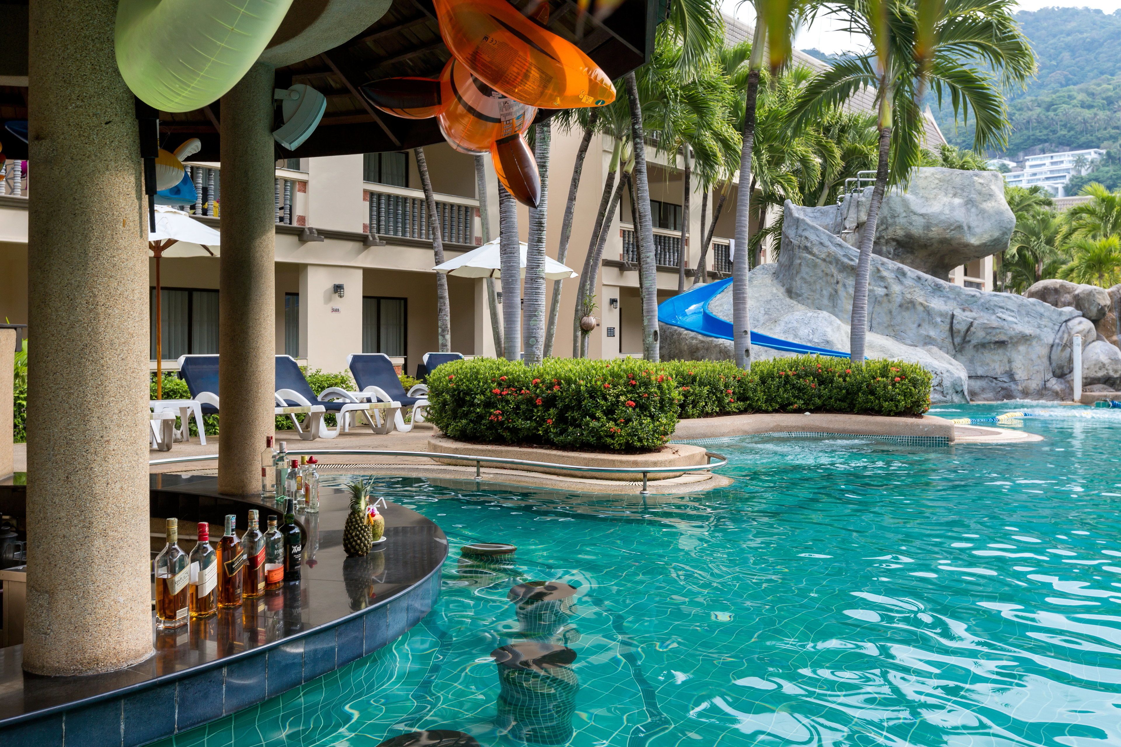 Un espace piscine avec des chaises longues, un bar bien garni et un décor tropical agrémenté de palmiers et d’une formation rocheuse.