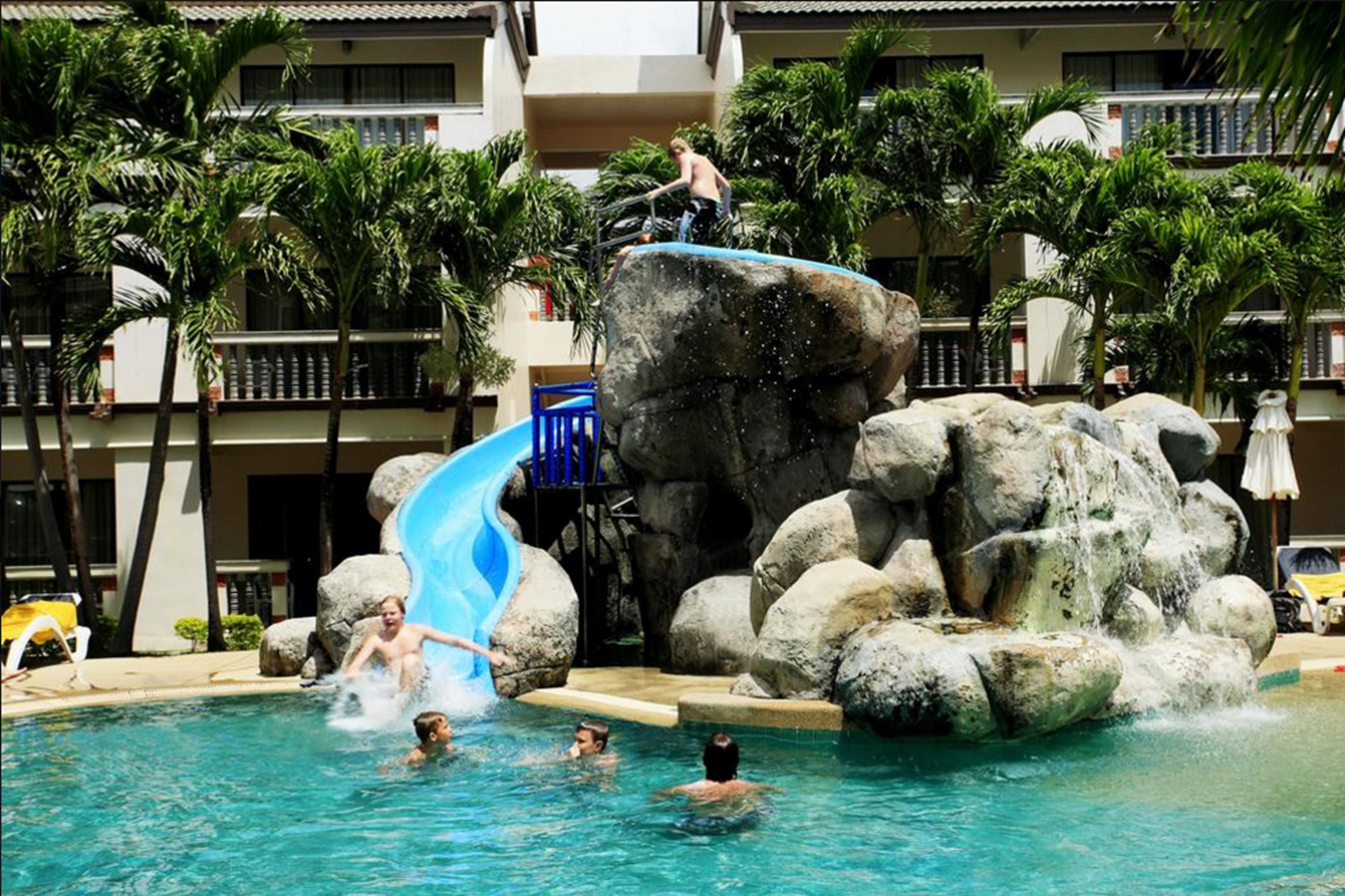 A pool area with a water slide, large rocks, and palm trees.