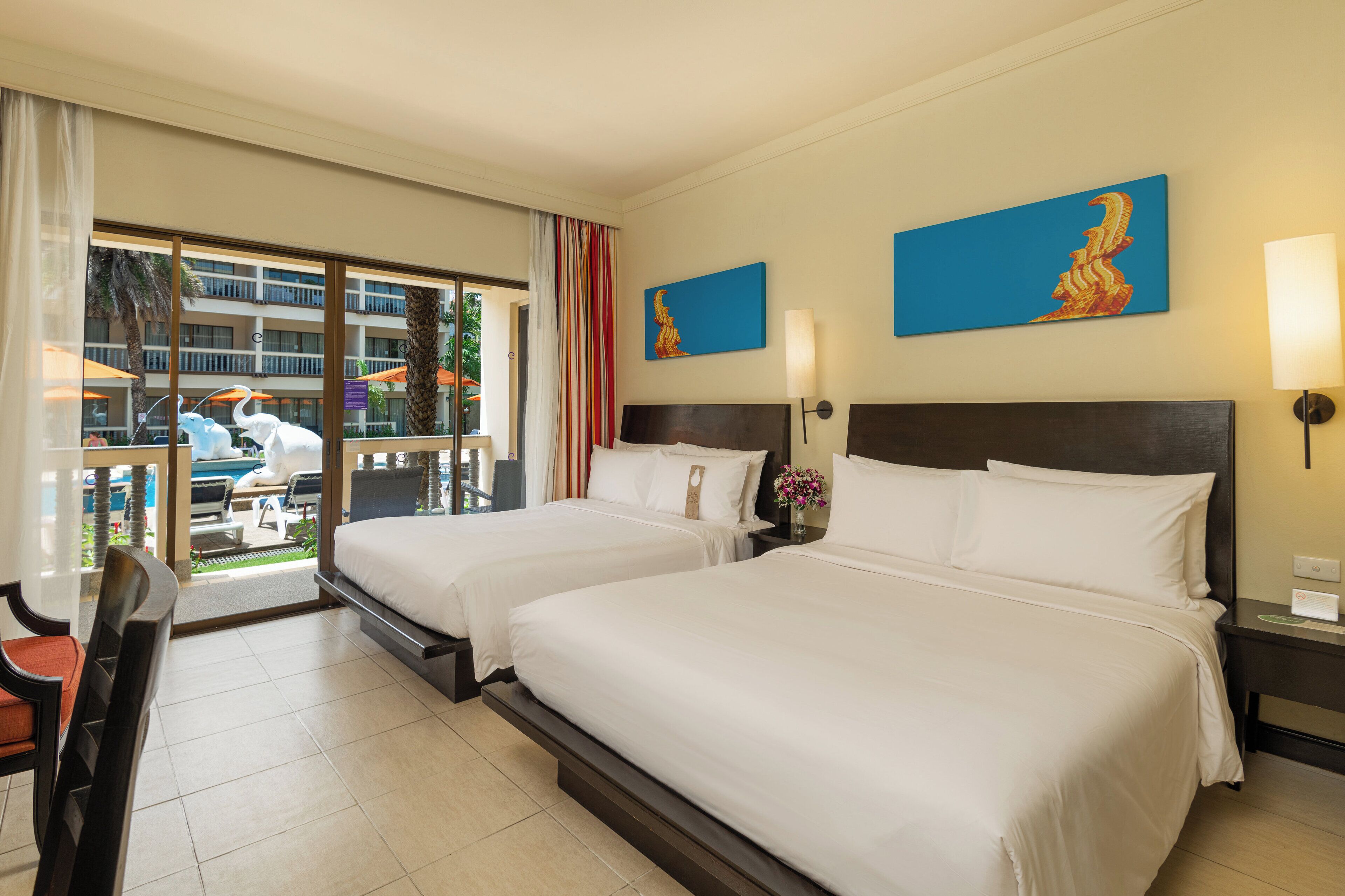 Une chambre d’hôtel avec deux lits, un balcon donnant sur la piscine et les palmiers, et deux tableaux accrochés au mur.