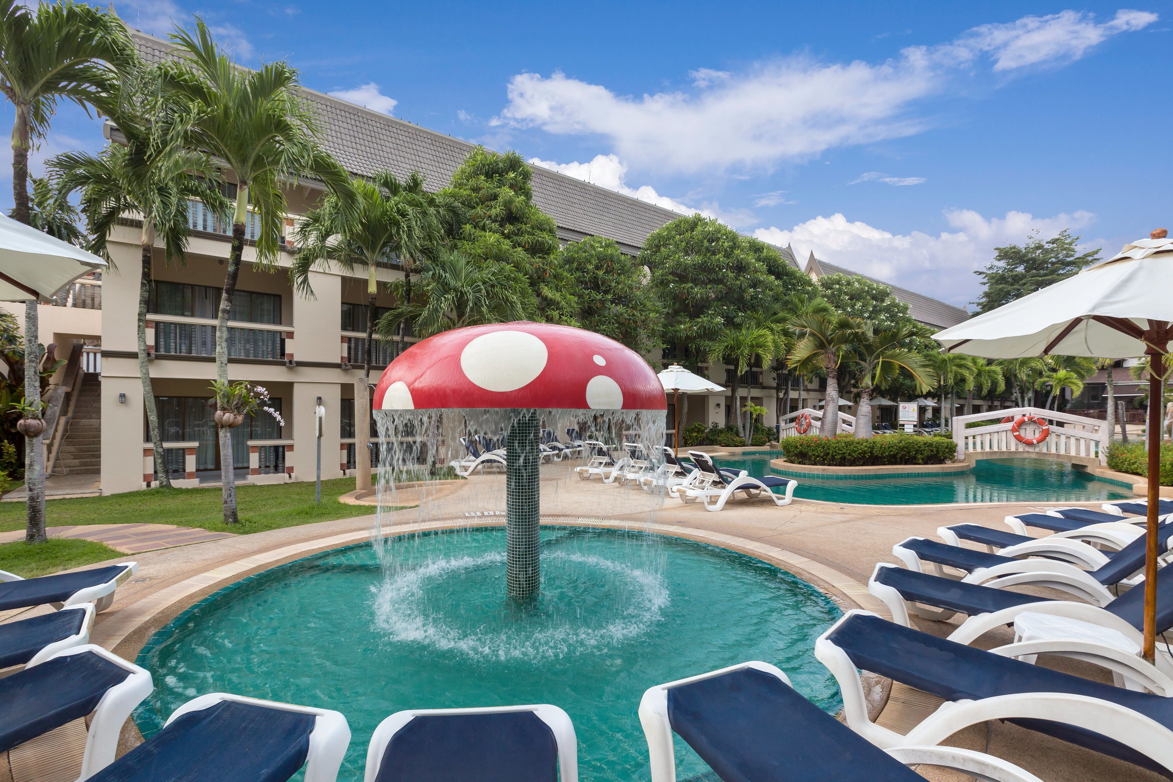 A pool area with lounge chairs, a water feature shaped like a mushroom, palm trees, and a building with a tiled roof.