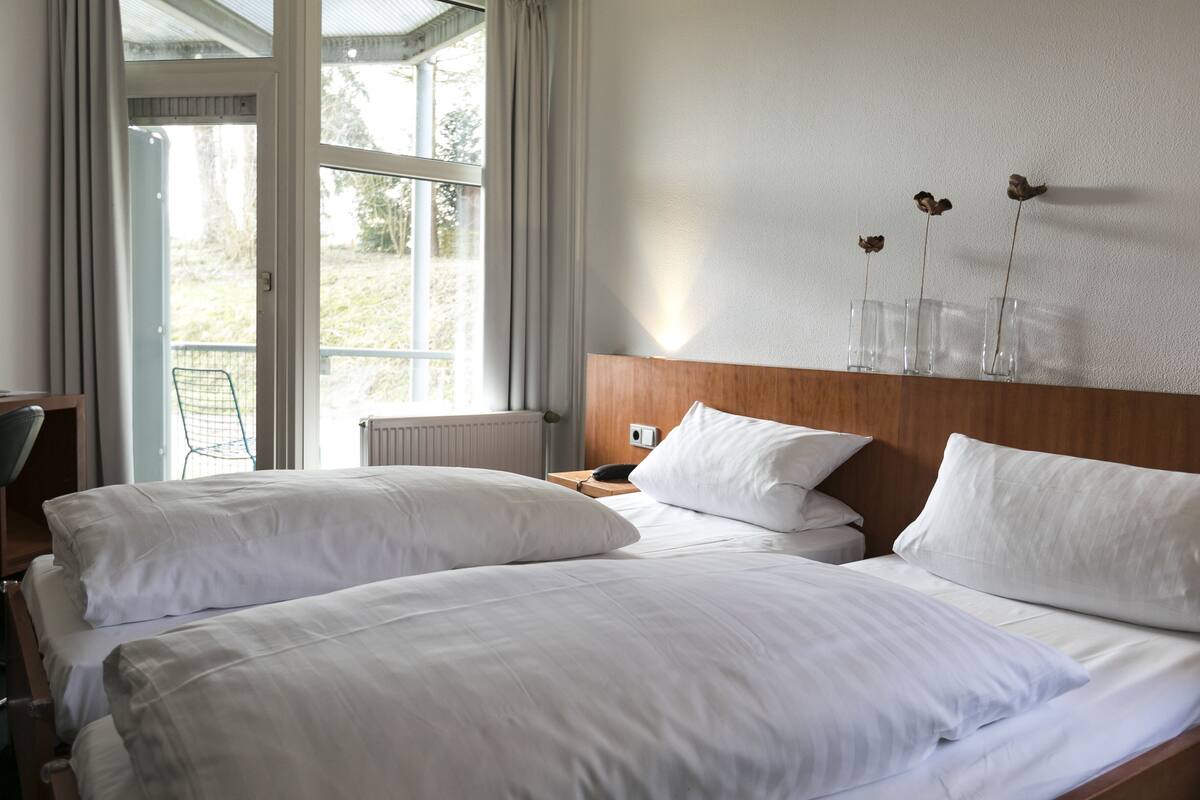 Een hotelkamer met twee bedden, een houten hoofdbord, een raam met gordijnen en een wandvaas met gedroogde bloemen.