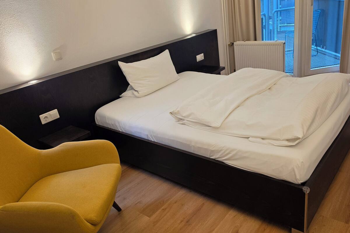 Een hotelkamer met een bed, een gele fauteuil, een nachtkastje, een raam met uitzicht en een radiator.
