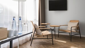 A modern room with a large window, a flat-screen TV, wicker chairs, a small round table, and a floor lamp. - Wald & Golfhotel Lottental (Bochum)