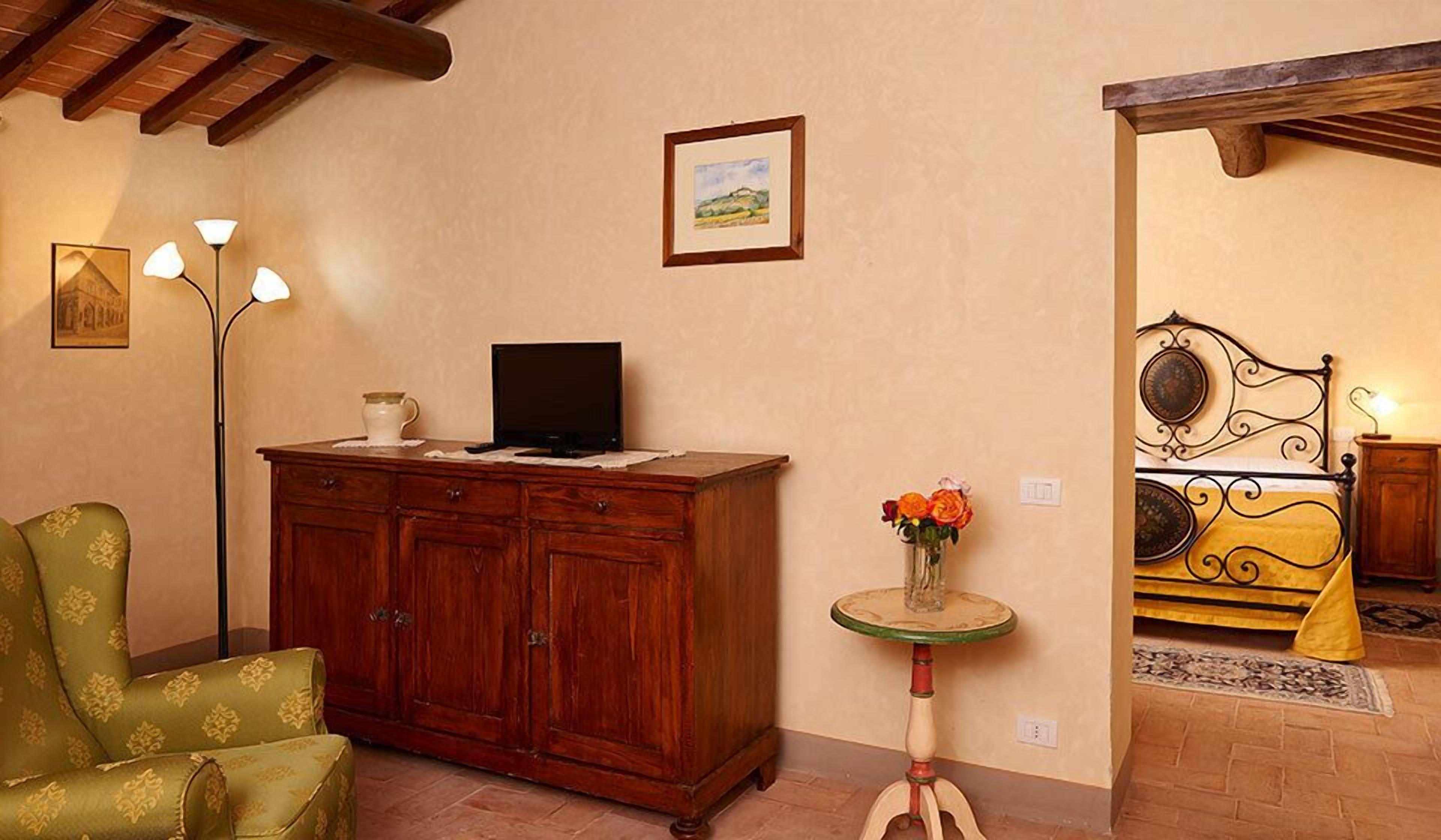 A room with a wooden cabinet, a television on top, a green armchair, a small table with a vase of flowers, and a bed with a metal frame.