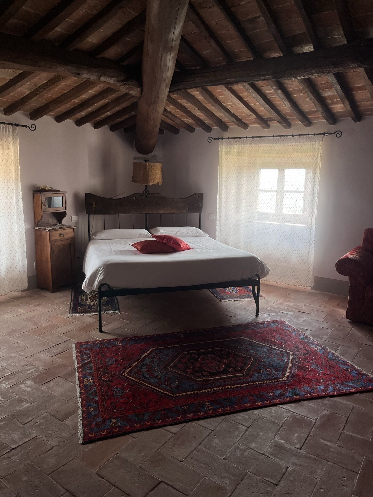A bedroom with a wooden ceiling, a bed with white linens and a red pillow, a wooden nightstand, a small table with a lamp, a patterned rug, and a window with sheer curtains.