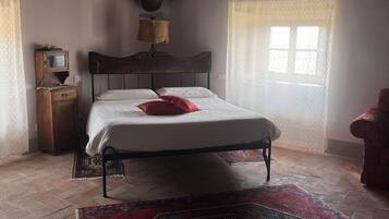 Una camera da letto con soffitto in legno, un letto con lenzuola bianche e un cuscino rosso, un comodino in legno, un tavolino con una lampada, un tappeto decorato e una finestra con tende leggere.
