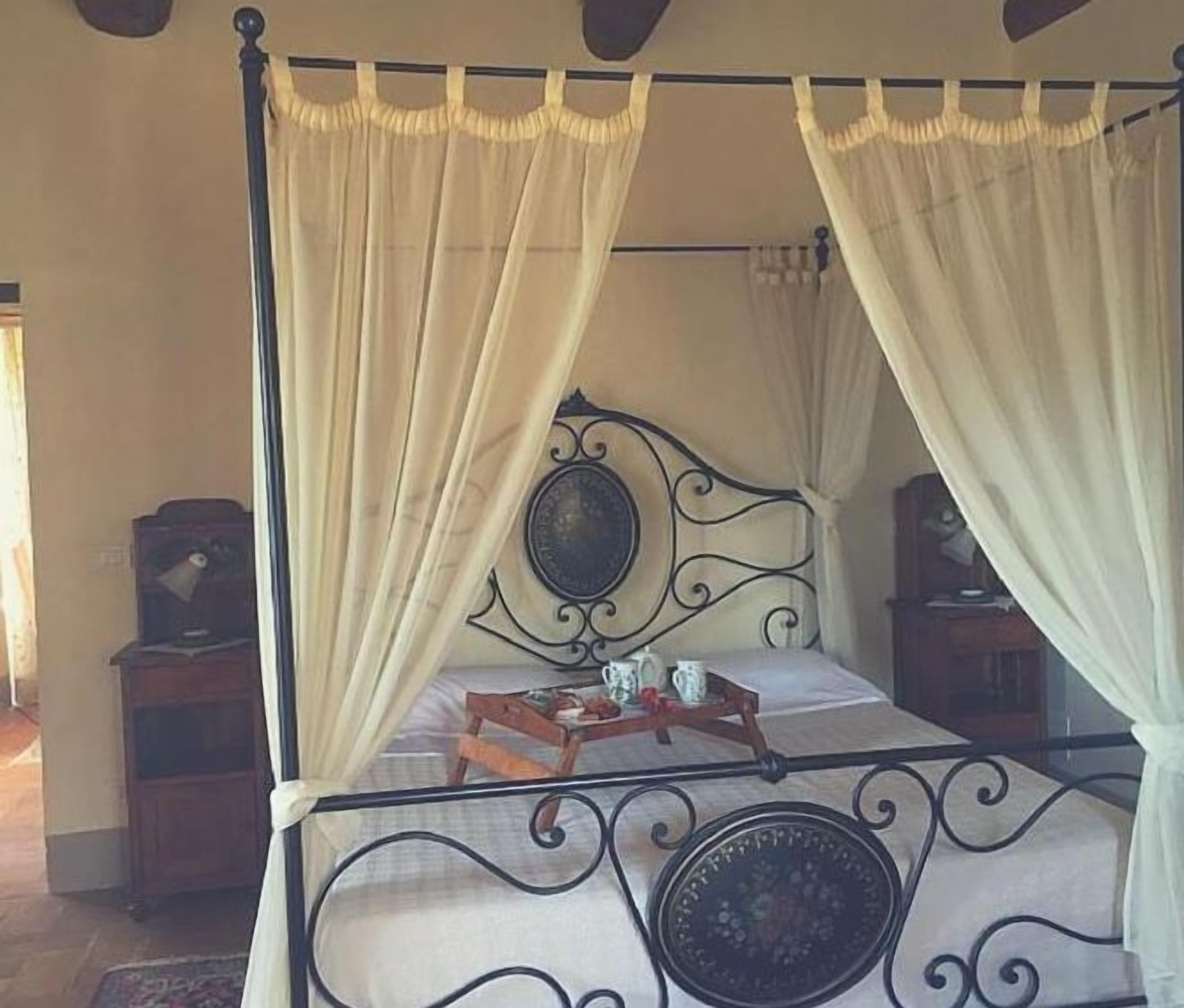 Una cama con dosel y cortinas, un espejo redondo sobre la cabecera y una mesita de noche con lámpara.