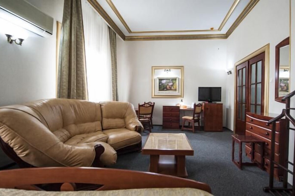 A room with a leather sofa, a wooden table, a desk with a chair, a TV, and a wardrobe.