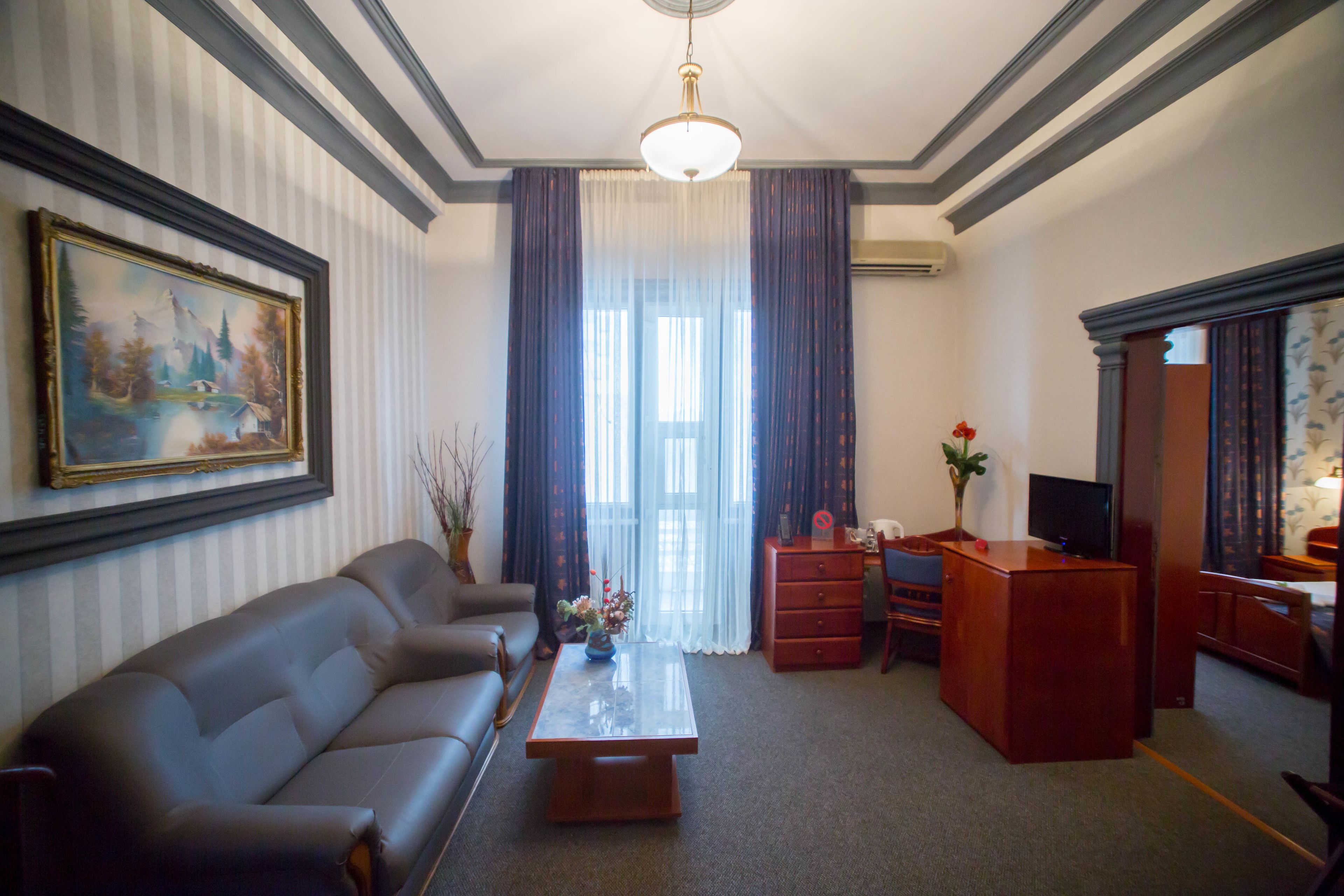 A hotel room with a leather sofa, a wooden coffee table, a desk with a television, and a large window with curtains.