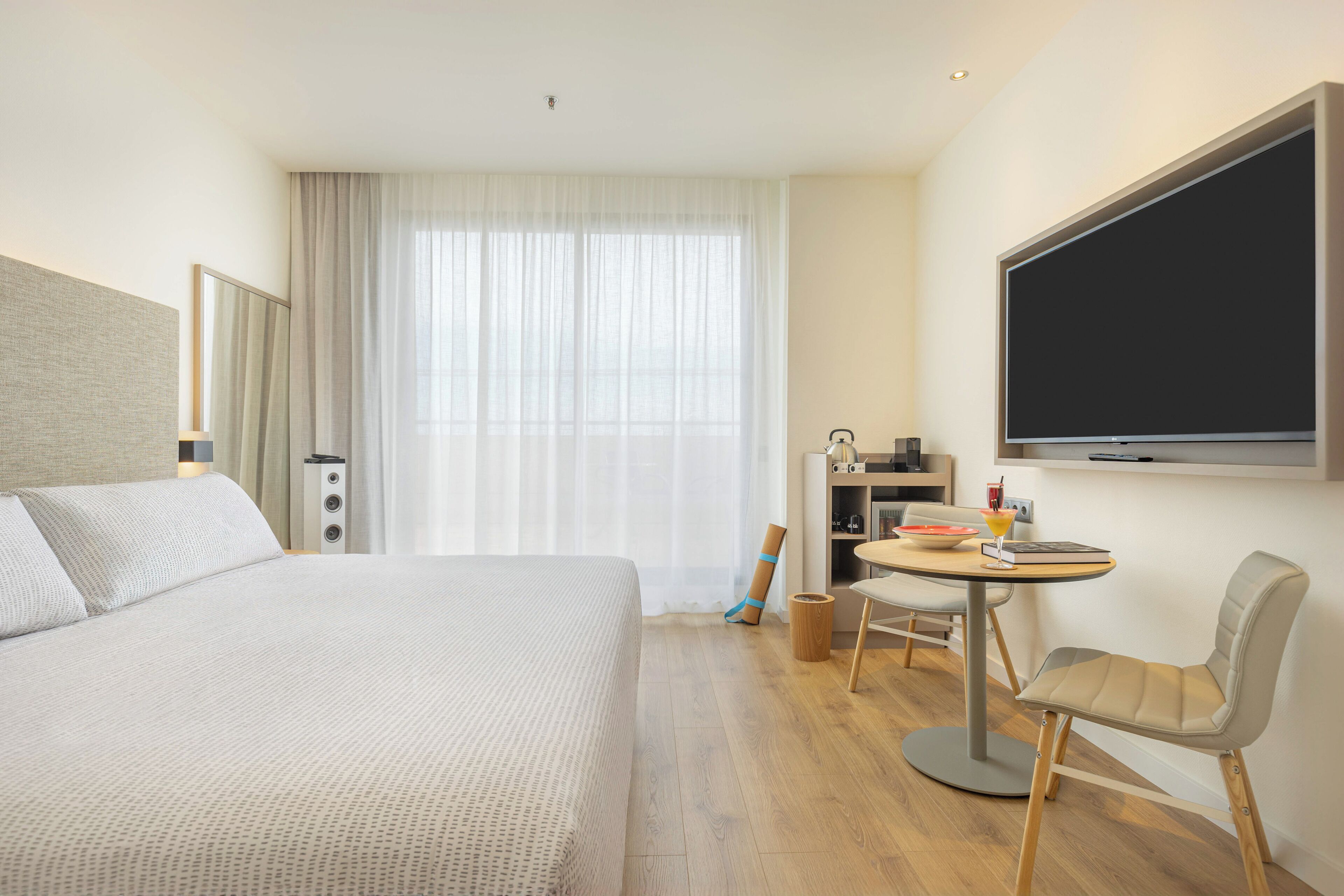 Quarto de hotel com cama, escrivaninha com cadeira, televisão e janela com cortinas.