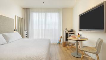 Quarto de hotel com cama, escrivaninha com cadeira, televisão e janela com cortinas.