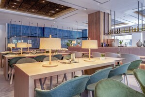 Un vestíbulo de hotel moderno con largas mesas de madera, sillas tapizadas de verde y una zona de bar con botellas.