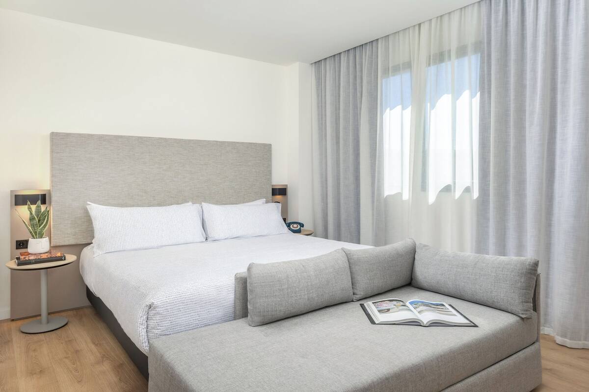 Una habitación de hotel moderna con una cama grande, un sofá gris, una mesita de noche con lámpara y una ventana con cortinas.