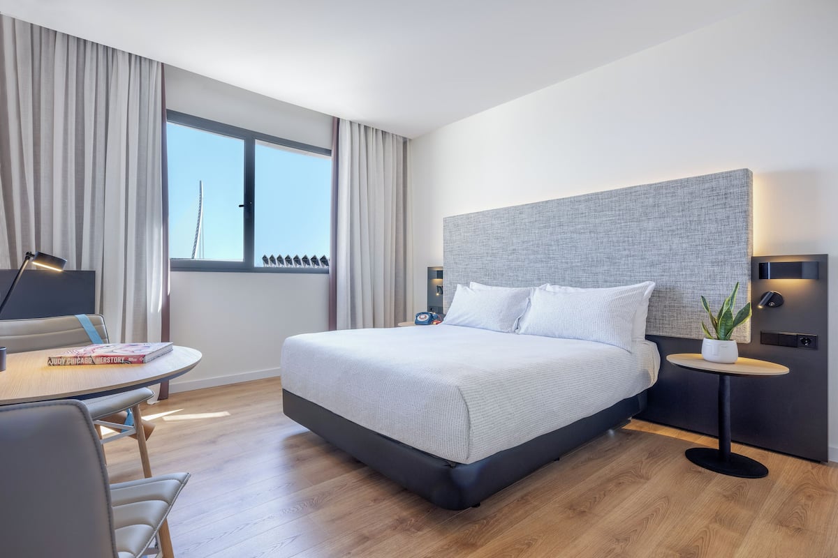 Una habitación de hotel moderna con una cama grande, una mesa redonda con una silla, una mesita auxiliar con una lámpara y vistas a la silueta urbana a través de la ventana.