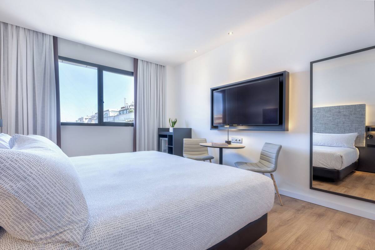 Habitación de hotel con una cama grande, un televisor montado en la pared, una mesita, dos sillas y un espejo amplio.