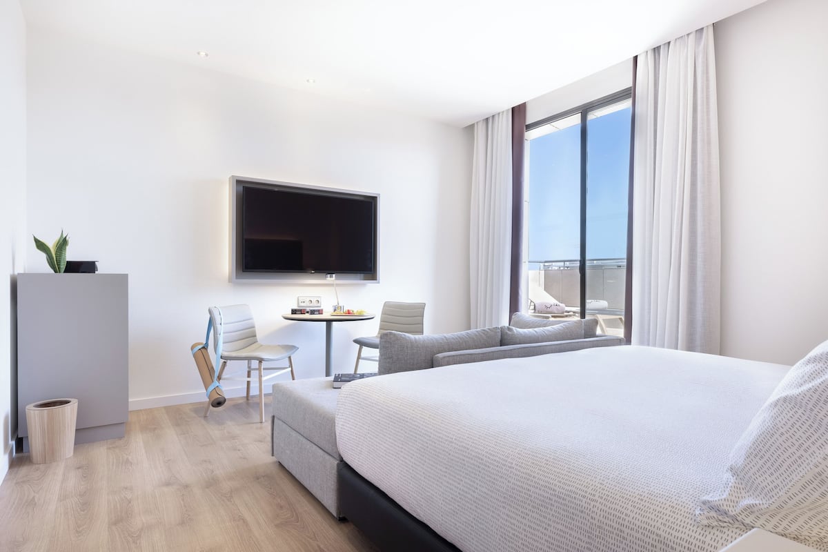 Una habitación de hotel moderna con una cama grande, un televisor de pantalla plana montado en la pared, una mesita redonda con una silla y un balcón con vistas al cielo.