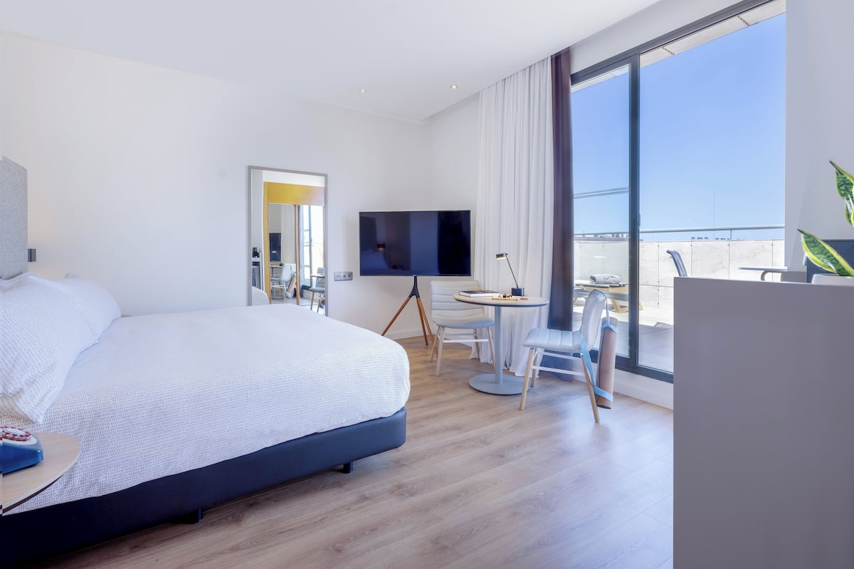 Una habitación de hotel moderna con una cama grande, televisión, escritorio con silla y balcón con vistas.