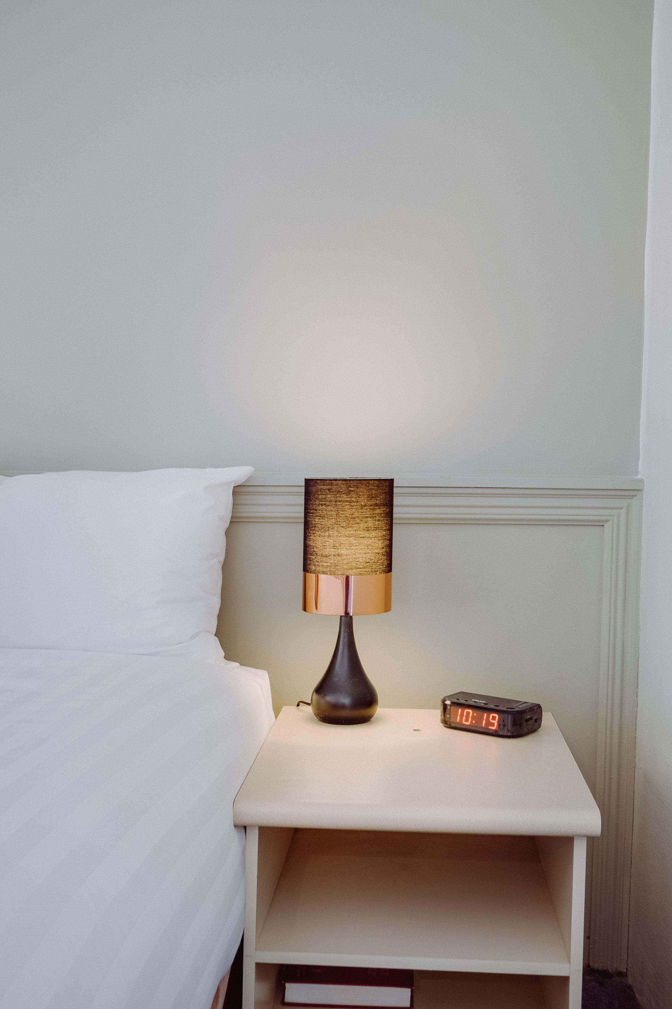 A neatly made bed with a white pillow and a white nightstand beside it. On the nightstand, there is a lamp with a textured lampshade and a digital clock displaying the time.