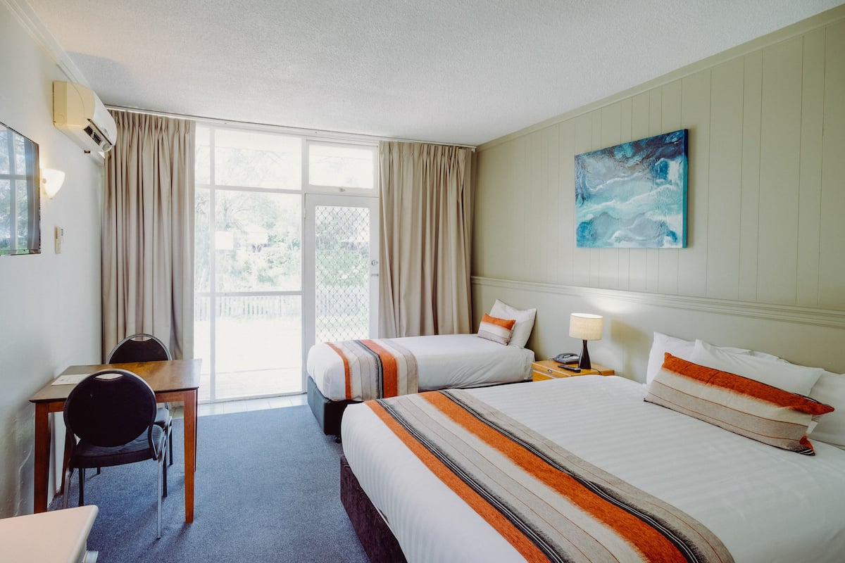 A hotel room with two beds, a desk, and a chair. There is a window with curtains, a painting on the wall, and a lamp on the nightstand.
