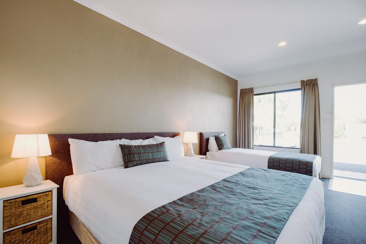 A hotel room with two beds, a nightstand with a lamp, a window with curtains, and a view of the outdoors.
