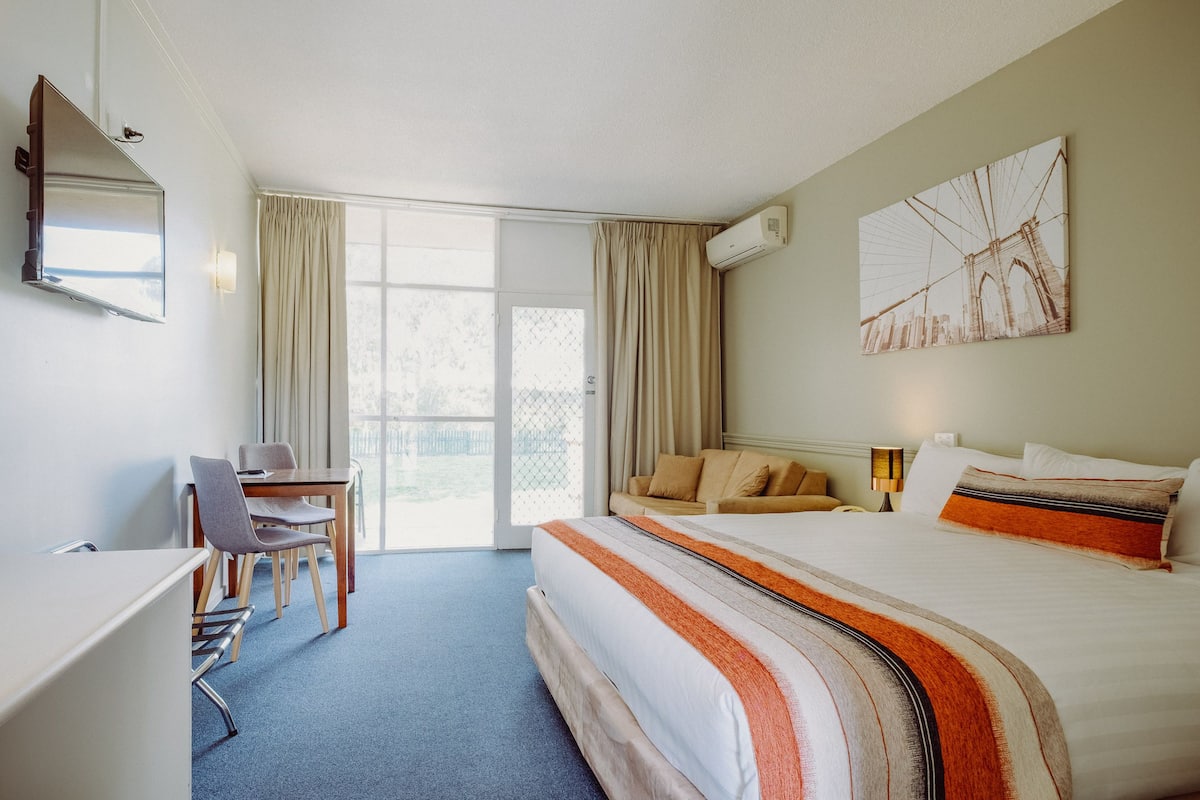 A hotel room with a bed, a desk, a chair, a large window, and a painting on the wall.