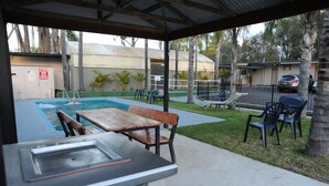 An outdoor pool area with a wooden table and chairs, a covered seating area, and a fenced parking lot. - Artesian Spa Motel (Moree)