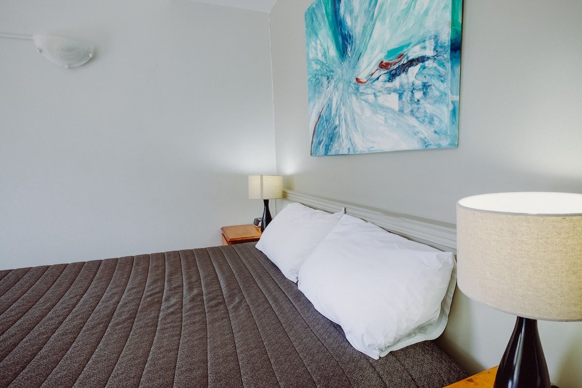 A neatly made bed with a brown quilt, a white pillow, and a white wall with a blue abstract painting.