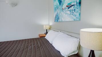 A neatly made bed with a brown quilt, a white pillow, and a white wall with a blue abstract painting.