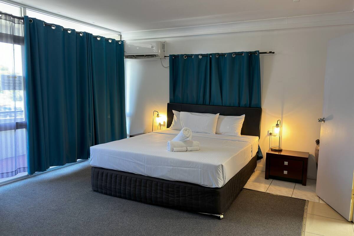 Une chambre d’hôtel avec un grand lit, des rideaux occultants, une fenêtre donnant sur un beau paysage et une table de chevet avec une lampe.