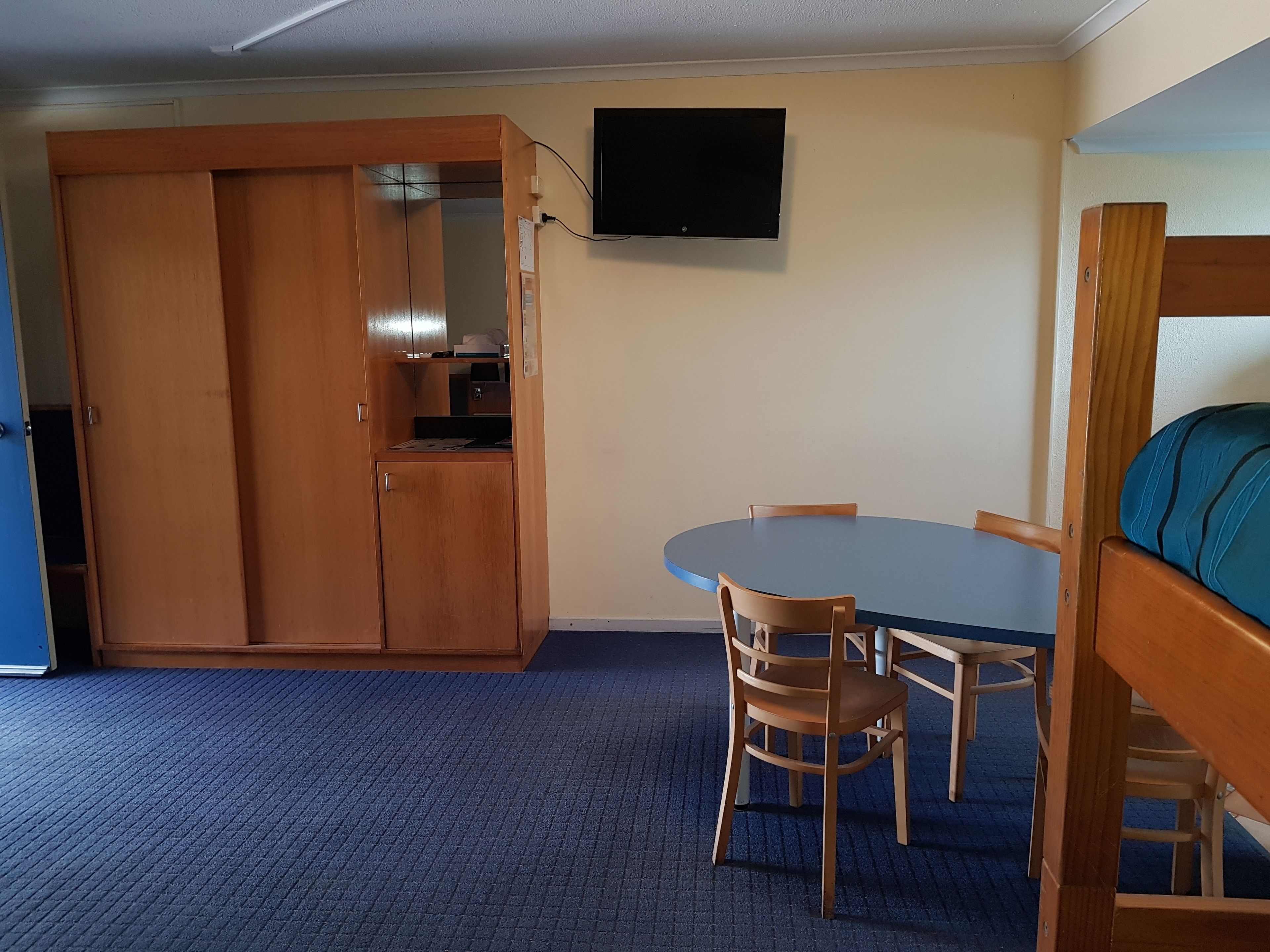 A room with a bunk bed, a dining table with chairs, a wooden wardrobe, and a television mounted on the wall.