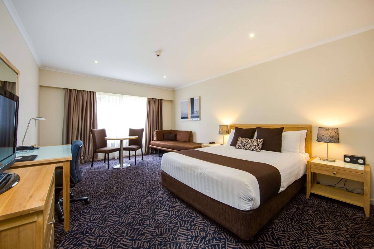 A hotel room with a large bed, a desk with a computer, a chair, a small table, and a TV.